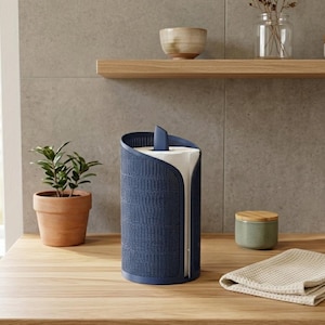 Pode incluir: Um porta-toalhas de papel azul marinho com uma textura tecida. O suporte tem um design curvo com uma ranhura para dispensar toalhas de papel. Uma pequena planta em vaso e uma toalha bege dobrada estão na superfície de madeira.