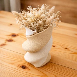 Op de afbeelding: Een decoratieve vaas met een uniek, gedraaid ontwerp, met een mix van beige en witte tinten. De vaas is gevuld met gedroogde bloemen en grassen in crème- en bruintinten, wat een natuurlijke, getextureerde esthetiek creëert. De vaas is ongeveer 25 cm hoog.