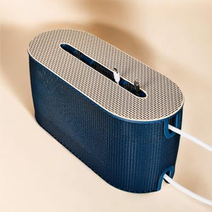 K&ouml;nnte beinhalten: Ein marineblauer Kabelorganizer mit ovaler Form und strukturierter Oberfl&auml;che. Die Oberseite ist beige mit einem gemusterten Design. Wei&szlig;e Kabel ragen aus den Seiten, und Ladekabel sind im Inneren sichtbar.