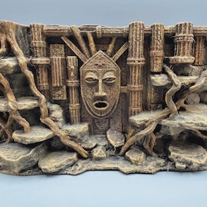 Pode incluir: Um ornamento retangular para aquário com uma máscara facial esculpida ao centro, rodeada por pilares texturizados semelhantes a pedra e raízes de árvore salientes. A estrutura inclui várias plataformas planas e em camadas com acabamento em bege e castanho.