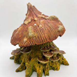 Puede incluir: Una escultura decorativa de un sombrero de hongo grande y texturizado de color marrón con manchas blancas, posado sobre una base de tocón de árbol de color amarillo verdoso cubierto de musgo y varios hongos pequeños en capas.