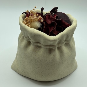 Pode incluir: Um recipiente de cerâmica bege texturizada em forma de saco, cheio de flores secas e potpourri bordô. O recipiente tem uma base arredondada e um topo franzido, criando uma peça decorativa rústica.