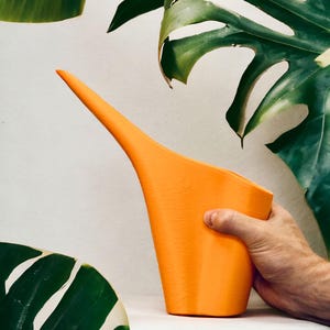 Pode incluir: Um regador laranja com um bico longo e fino é segurado por uma mão. O regador é feito de um material sólido e opaco. Grandes folhas verdes de uma planta estão ao fundo.