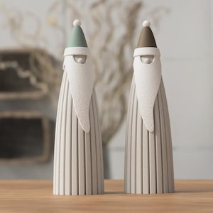 Pode incluir: Duas figuras cónicas altas com corpos cinzentos claros canelados. Cada figura apresenta uma barba branca texturizada e um chapéu pontiagudo, um em verde sálvia suave e o outro em castanho taupe, ambos com uma pequena esfera branca.