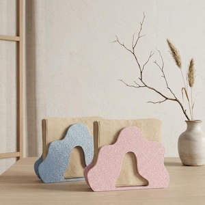 Pode incluir: Dois suportes decorativos para sacos de papel em forma de arco, um azul claro e outro rosa, ambos com acabamento brilhante. Estão sobre uma mesa de madeira, segurando sacos de papel castanhos. Um vaso com plantas secas ao fundo.
