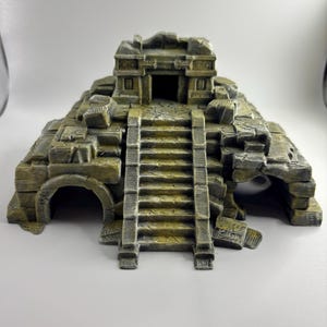 May include: A decorative aquarium ornament shaped like an ancient stone temple ruin. The structure features a central staircase leading to an elevated doorway, flanked by two arched side openings, all finished in a weathered gray and yellow stone texture.