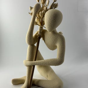Pode incluir: Uma escultura bege de uma figura sentada segurando um buquê de flores secas e um bastão de madeira. A figura está sentada, com os braços enrolados nas pernas. As flores são de cor marrom claro e o bastão é marrom escuro.
