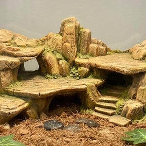 May include: A detailed, multi-level terrarium decoration resembling a rock formation with a cave, steps, and platforms. The structure is primarily tan and brown with green accents, and is designed for reptiles or small animals.