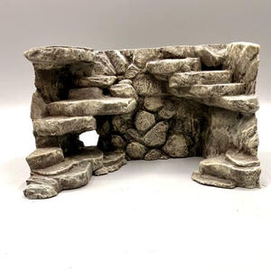 Pode incluir: Uma estrutura rochosa em tons de cinza e bege com múltiplas plataformas e saliências em camadas. A superfície tem uma aparência texturizada semelhante a pedra, com detalhes esculpidos que lembram formações rochosas naturais e uma abertura.