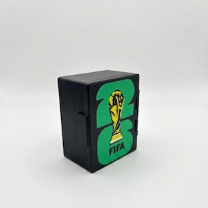 May include: A rectangular black plastic storage box featuring a green graphic of the number 26 with the FIFA World Cup trophy silhouette and the text FIFA printed in black. The box has a hinged side design and a textured matte finish surface.