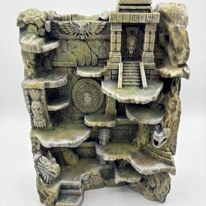 Pode incluir: Um expositor com textura de pedra em tons de bege e verde, com design de templo antigo, apresentando múltiplas plataformas em camadas, relevos esculpidos de rostos, um motivo central de calendário circular e uma pequena escada de acesso.
