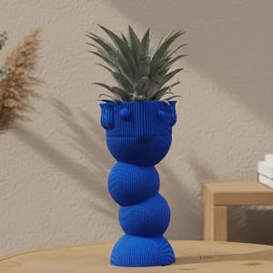 Può includere: Un vaso decorativo blu con un design a forma di volto, che contiene una pianta verde. Il vaso ha un design a sfere impilate ed è posto su un tavolo di legno. A sinistra c'è un vaso bianco con piante essiccate.