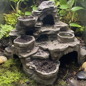 May include: A gray, tiered terrarium decoration resembling a rock formation with multiple planters filled with soil. The structure has a cave opening and small water basins. Green plants and moss surround the decoration, with small stones scattered around.