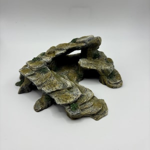 Pode incluir: Um ornamento de formação rochosa texturizada com superfícies de pedra em camadas em tons de bege, castanho e cinzento com manchas de musgo verde. A estrutura inclui uma abertura semelhante a uma gruta e saliências em degraus.