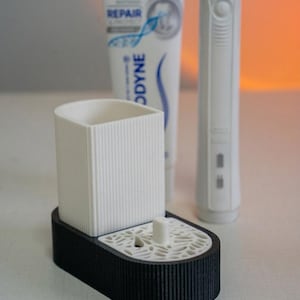 May include: A white and black toothbrush holder with a ribbed design, holding a toothbrush and toothpaste. The toothpaste tube is white with blue and green accents, and the text "REPAIR" is visible.