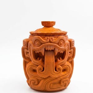 May include: A terracotta-colored lidded jar featuring a relief sculpture of a stylized creature with a wide open mouth, prominent fangs, and a long protruding tongue. The surface is covered in intricate geometric patterns and carvings.