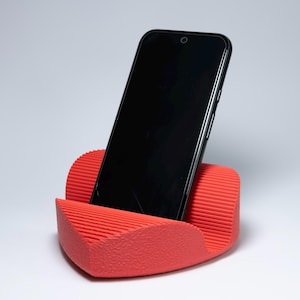 Pode incluir: Um suporte de telefone vermelho com um design texturizado e canelado, segurando um smartphone preto. O suporte é inclinado para suportar o telefone, em um fundo branco.