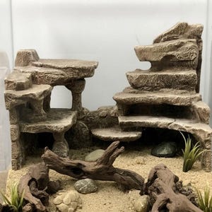 May include: A detailed aquarium decoration featuring a layered rock formation with a naturalistic appearance. The scene includes sand, small rocks, driftwood, and artificial plants, creating a miniature desert landscape for aquatic pets.