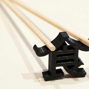 Pode incluir: Um descanso para pauzinhos preto em forma de caractere Kanji japonês, segurando dois pauzinhos de madeira. O descanso tem um design geométrico e está sobre um fundo branco.