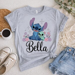 May include: A light gray t-shirt featuring a cartoon graphic of a blue alien character and a green alien character, surrounded by pink and purple flowers. The name "Bella" is written in a cursive font below the graphic.