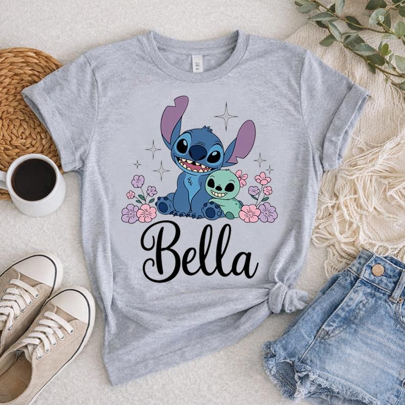 May include: A light gray t-shirt featuring a cartoon graphic of a blue alien character and a green alien character, surrounded by pink and purple flowers. The name "Bella" is written in a cursive font below the graphic.