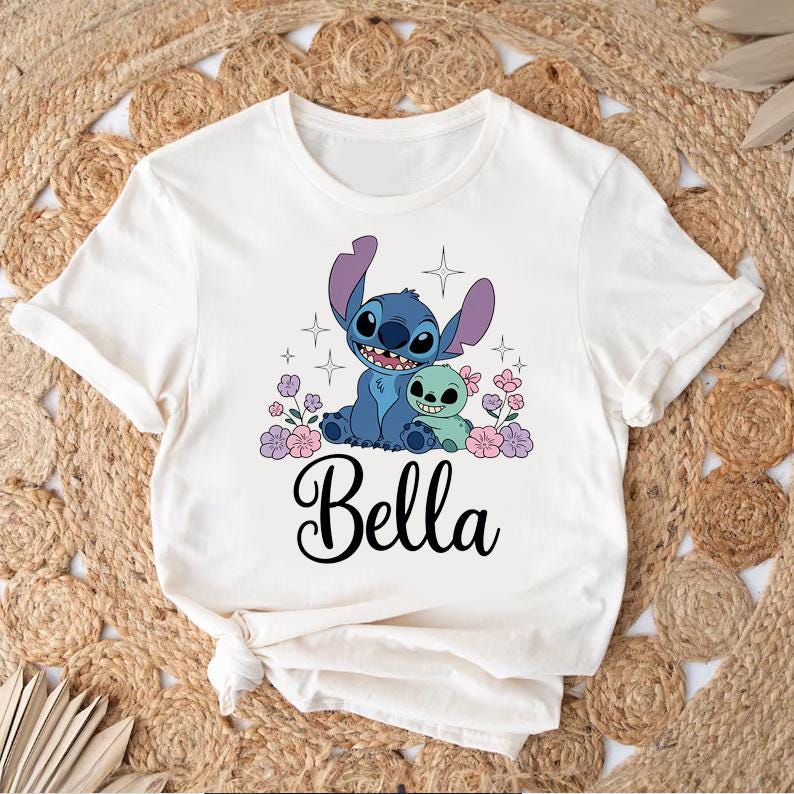 May include: White t-shirt featuring a graphic of two cartoon characters, one blue and one green, surrounded by flowers and stars. The name "Bella" is written in cursive below the characters.