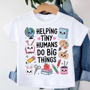 Puede incluir: Camiseta blanca con el texto "Helping Tiny Humans Do Big Things" y varias ilustraciones coloridas de útiles escolares. El diseño incluye un globo terráqueo, un libro, una paleta de pintura, un lápiz y otros gráficos lindos.