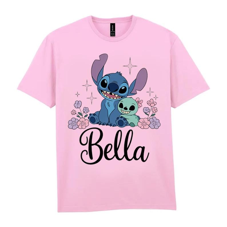 May include: A light pink t-shirt featuring a blue cartoon character with large ears and a smaller green character. The word "Bella" is written in cursive below the characters, surrounded by flowers and stars.