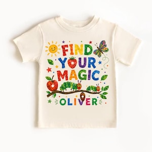 以下が含まれることがあります： 「FIND YOUR MAGIC OLIVER」というテキストが書かれたカラフルなグラフィックが特徴の、クリーム色の半袖Tシャツ。デザインには、黄色い太陽、イチゴ、蝶、赤いリンゴ、枝に乗った緑色の毛虫、そして繭が描かれています。