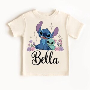 May include: Cream-colored t-shirt featuring a blue cartoon character with large ears and a smaller green character. The design includes pink and purple flowers, stars, and the name "Bella" in black script.