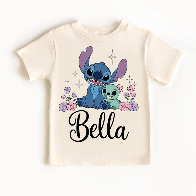 May include: Cream-colored t-shirt featuring a blue cartoon character with large ears and a smaller green character. The design includes pink and purple flowers, stars, and the name "Bella" in black script.