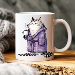 Könnte beinhalten: Weiße Keramiktasse mit einer Cartoon-Katze in einem lila karierten Bademantel, die eine dampfende Kaffeetasse hält. Die Katze hat einen entspannten Ausdruck, mit Kaffeebohnen zu ihren Füßen. Die Tasse hat einen gebogenen Griff.