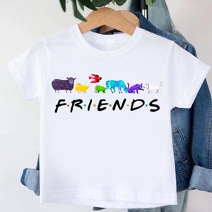 May include: White t-shirt featuring the word "FRIENDS" in black capital letters, with colorful animal illustrations replacing the letters in the word. The animals include a sheep, duck, bird, frog, horse, cat, and dog.