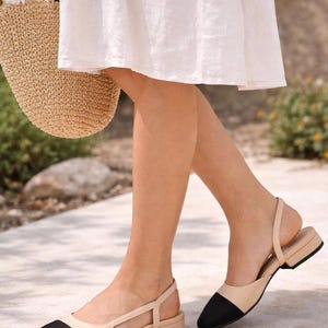 K&ouml;nnte beinhalten: Beige und schwarze Slingback-Flats mit kleinem Absatz. Die Schuhe haben eine spitze Zehenpartie und einen Kn&ouml;chelriemen. Die Schuhe werden im Freien getragen.