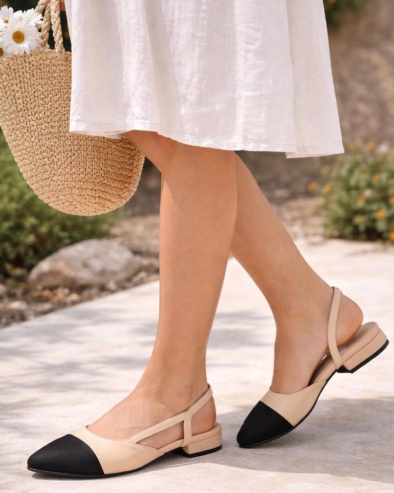 K&ouml;nnte beinhalten: Beige und schwarze Slingback-Flats mit kleinem Absatz. Die Schuhe haben eine spitze Zehenpartie und einen Kn&ouml;chelriemen. Die Schuhe werden im Freien getragen.