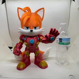 May include: A 3D-printed figurine of Tails from Sonic the Hedgehog, reimagined in an Iron Man suit. The figure is predominantly orange, red, and gold, with a stern expression. A clear plastic water bottle is next to the figure.
