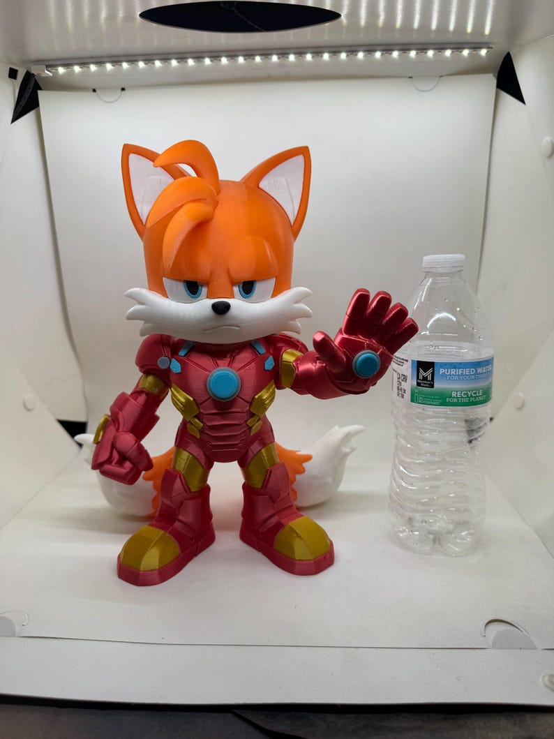 May include: A 3D-printed figurine of Tails from Sonic the Hedgehog, reimagined in an Iron Man suit. The figure is predominantly orange, red, and gold, with a stern expression. A clear plastic water bottle is next to the figure.