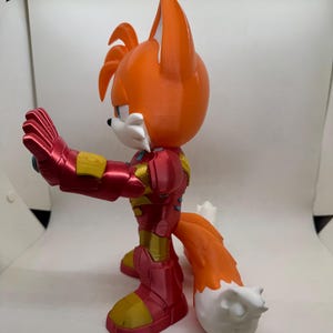 May include: A 3D-printed figurine of Tails from Sonic the Hedgehog, reimagined in an Iron Man-inspired suit. The figure features a vibrant orange head and tail with white accents, and a red and gold armored body. The figure is posed with one arm extended.