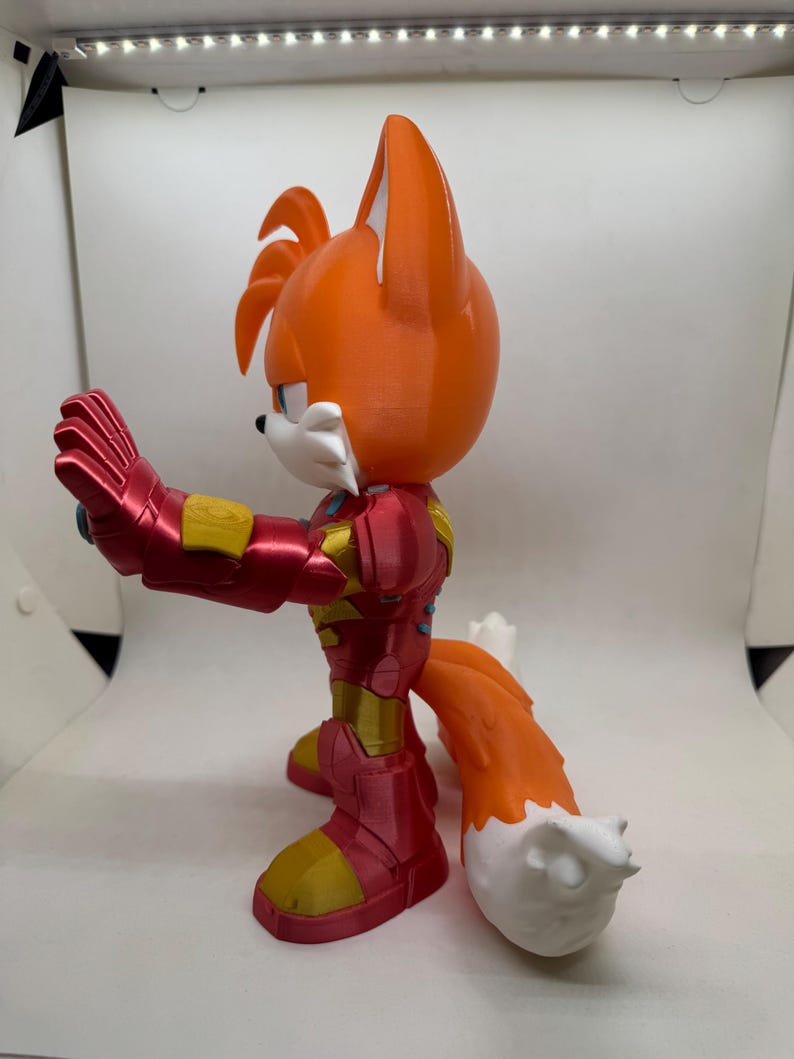 May include: A 3D-printed figurine of Tails from Sonic the Hedgehog, reimagined in an Iron Man-inspired suit. The figure features a vibrant orange head and tail with white accents, and a red and gold armored body. The figure is posed with one arm extended.