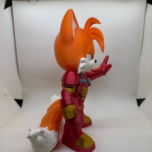 May include: A 3D-printed figurine of Tails from Sonic the Hedgehog, wearing a red and gold Iron Man suit. The figure has orange fur, white accents, and is posed with one hand raised. The figure is on a white background.