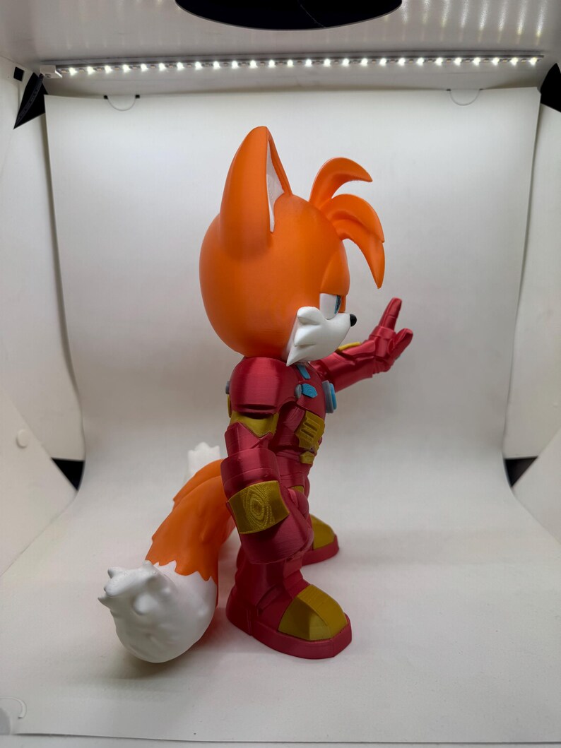 May include: A 3D-printed figurine of Tails from Sonic the Hedgehog, wearing a red and gold Iron Man suit. The figure has orange fur, white accents, and is posed with one hand raised. The figure is on a white background.