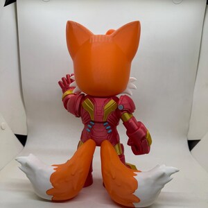 May include: A stylized orange fox figure with white accents and a red and gold armored suit. The figure has large ears and a waving hand. The fox's tail is orange with white tips. The figure is standing on a white surface.