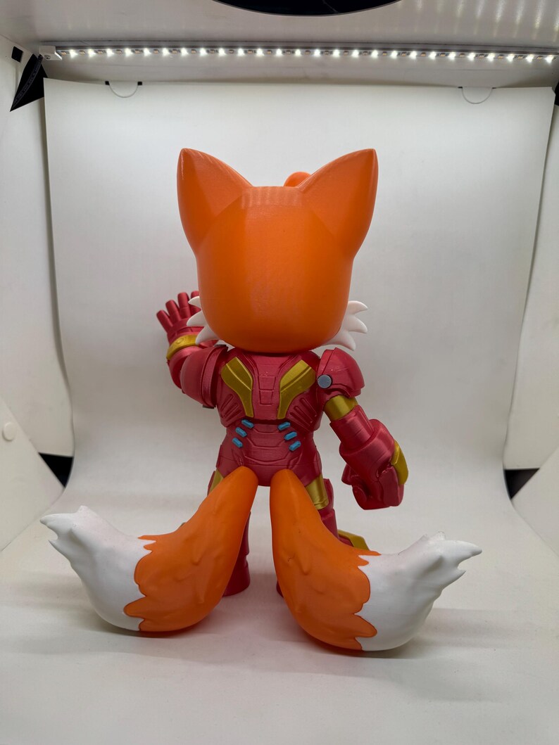 May include: A stylized orange fox figure with white accents and a red and gold armored suit. The figure has large ears and a waving hand. The fox's tail is orange with white tips. The figure is standing on a white surface.