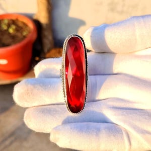 May include: A silver ring featuring a large, faceted, oval-shaped, deep red gemstone. The gemstone is set in a silver band with a rope-like border. The ring is displayed on a white surface.