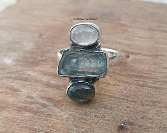 May include: A silver ring featuring three gemstones: a clear, oval-shaped stone at the top, a rectangular, light blue stone in the center, and a dark, oval-shaped stone at the bottom. The ring is set on a wooden surface.
