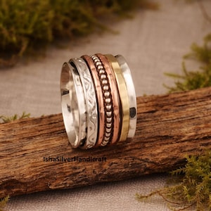 以下が含まれることがあります： シルバー、銅、ゴールドのデザインが施されたマルチバンドリング。 リングは、テクスチャ加工とビーズ加工が施されたバンドが特徴です。 ブランド名「IshaSilverHandicraft」が木の表面に表示されています。