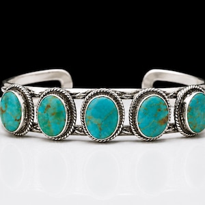 Könnte beinhalten: Ein Silberarmreif mit fünf ovalen Türkissteinen, die braune Matrixmuster aufweisen. Jeder Stein ist in eine gedrehte Kordelbordüre gefasst, und das Armband besteht aus zwei parallelen Silberdrähten mit dekorativen, eingravierten Details.
