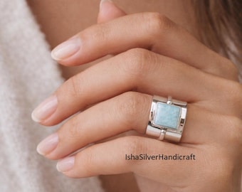 Può includere: Un anello d'argento con una pietra preziosa rettangolare azzurra. L'anello ha una fascia larga con un design moderno. La pietra preziosa è incastonata in una cornice quadrata. L'anello è su una mano.