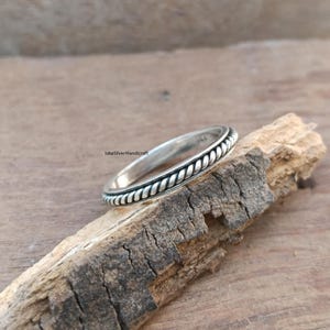 May include: A silver ring featuring a braided rope pattern around the circumference. The ring is displayed on a piece of natural wood. A simple and elegant piece of jewellery.