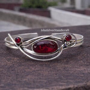 May include: A silver cuff bracelet with three red gemstones. The central stone is oval-shaped, flanked by two smaller, round stones. The bracelet has a delicate, swirling design and is set against a textured background. The text "IshaSilverHandicraft" is visible.
